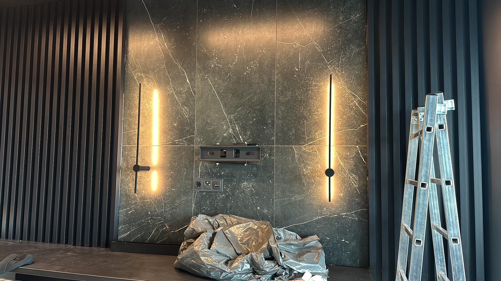Ściana z marmurową okładziną i podświetleniem LED, widoczny uchwyt na telewizor i gniazdka elektryczne, zabezpieczone przed zabrudzeniem, drabina aluminiowa po prawej stronie.