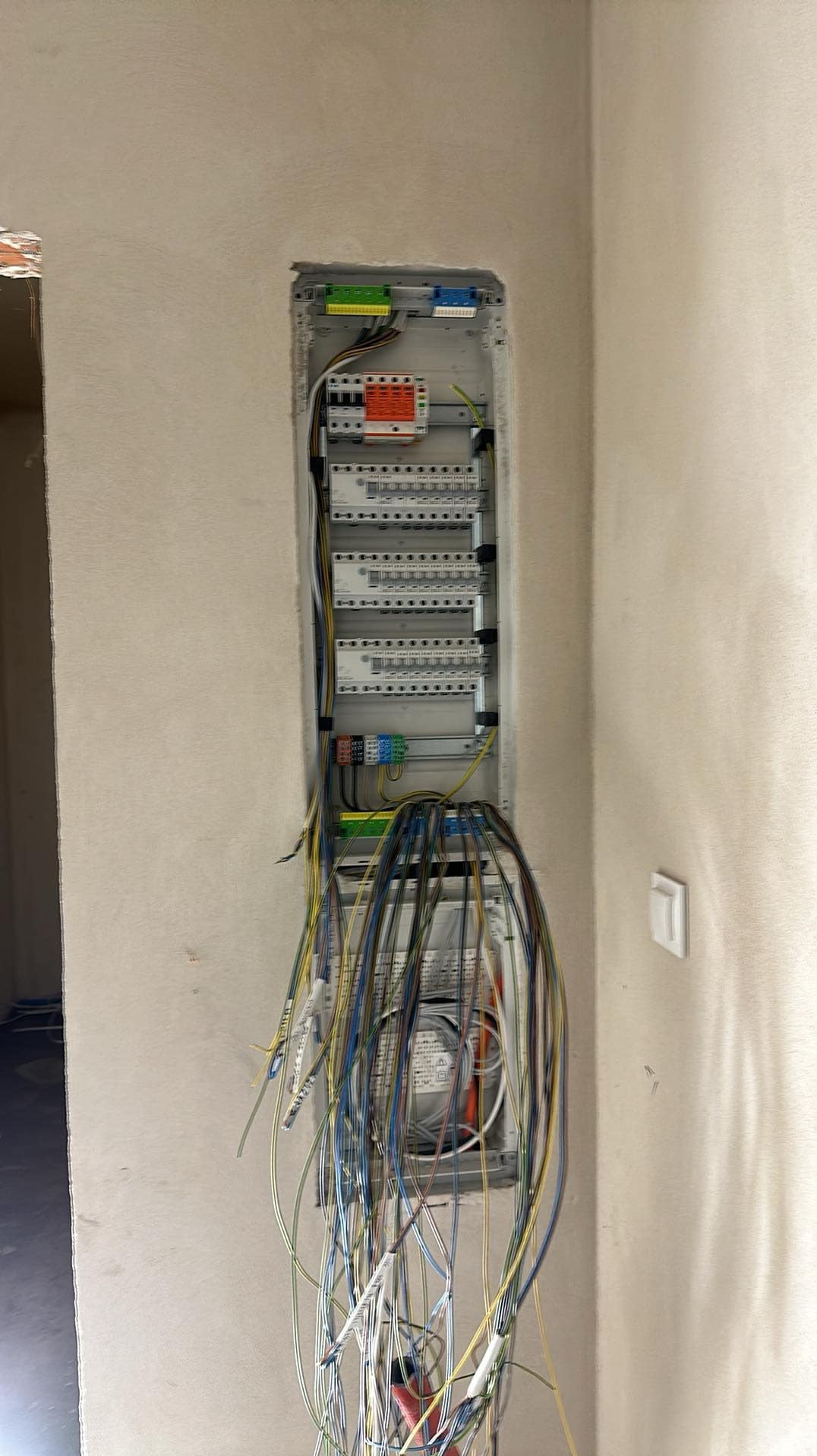 Szafka rozdzielcza w ścianie w trakcie montażu, widoczne liczne przewody elektryczne z oznaczeniami, w tle surowa ściana i fragment otworu drzwiowego.