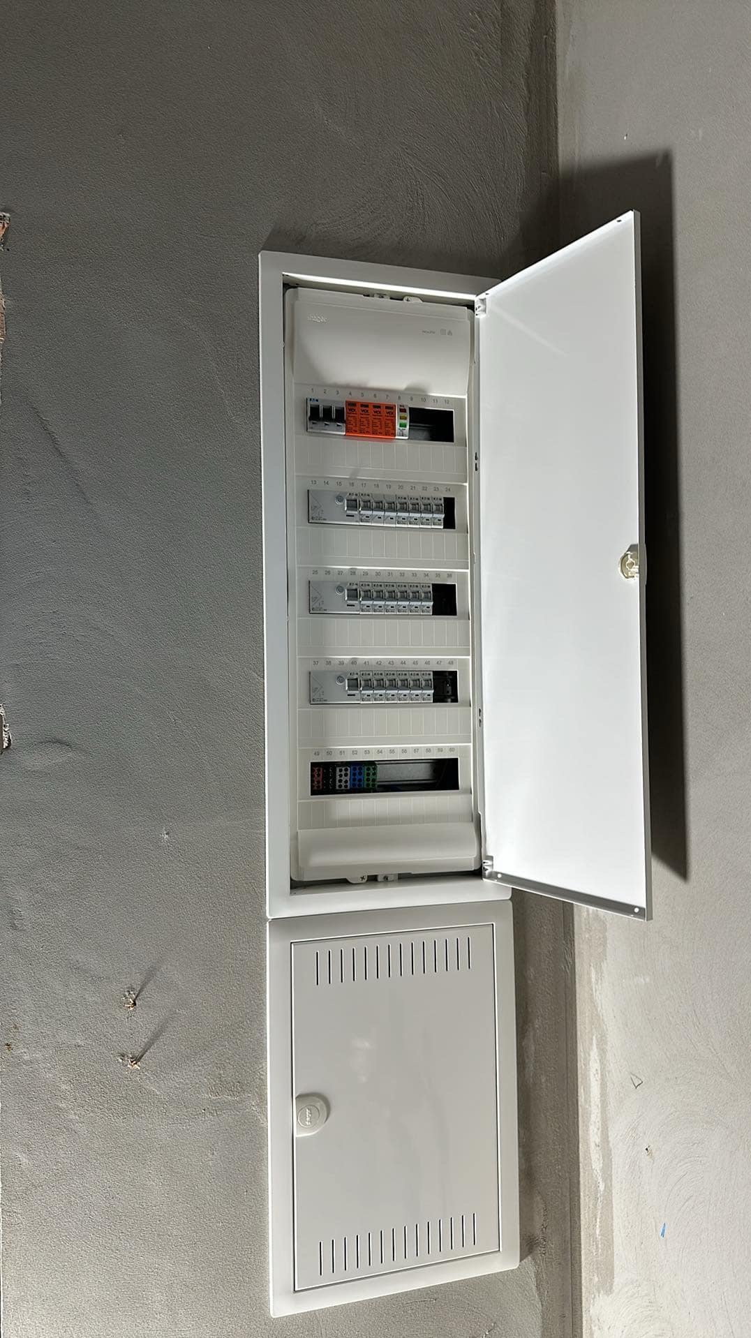 Otwarta, biała skrzynka rozdzielcza z bezpiecznikami i wyłącznikami zamontowana na surowej, szarej ścianie. Widoczne numery obwodów. Dolna część skrzynki zamknięta, z perforowaną obudową.