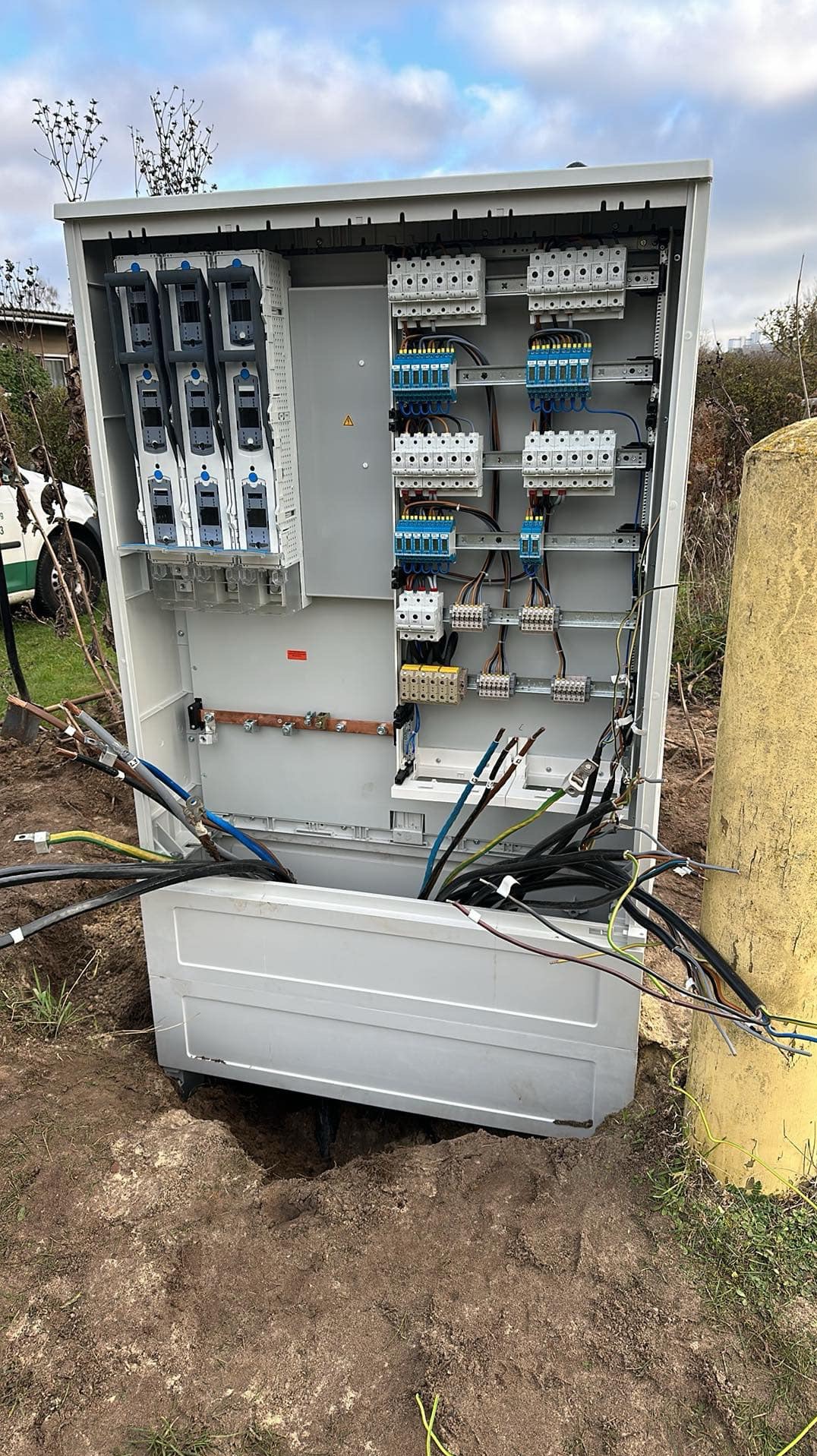 Otwarta skrzynka elektryczna na zewnątrz z widocznymi bezpiecznikami, złączkami i kablami w ziemi, obok żółtego słupa.