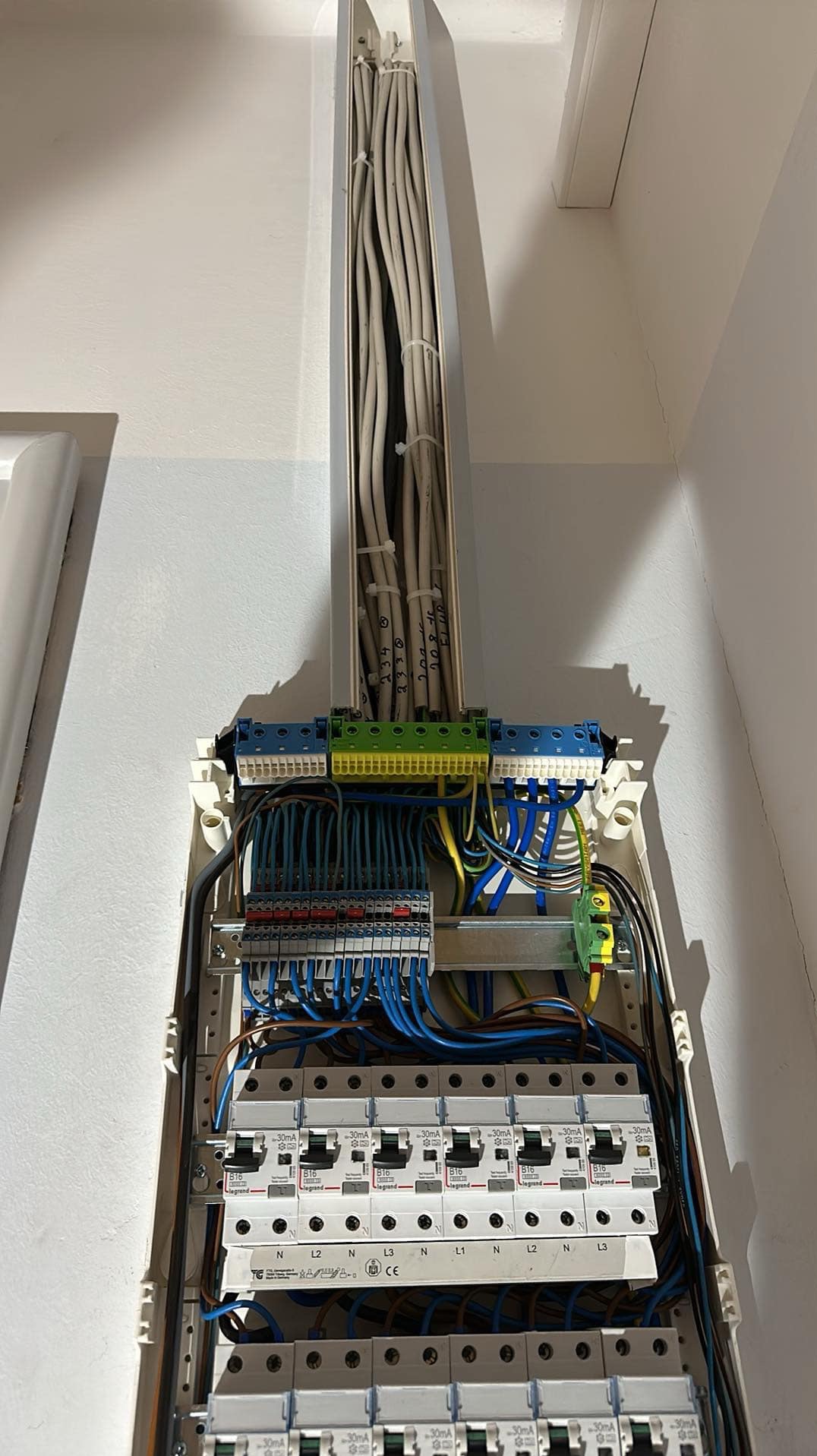 Rozdzielnica elektryczna z licznymi przewodami w białej obudowie, widoczne zabezpieczenia topikowe i różnicowoprądowe marki Legrand, kable prowadzone w korytku pionowo do góry z numeracją na kablach.