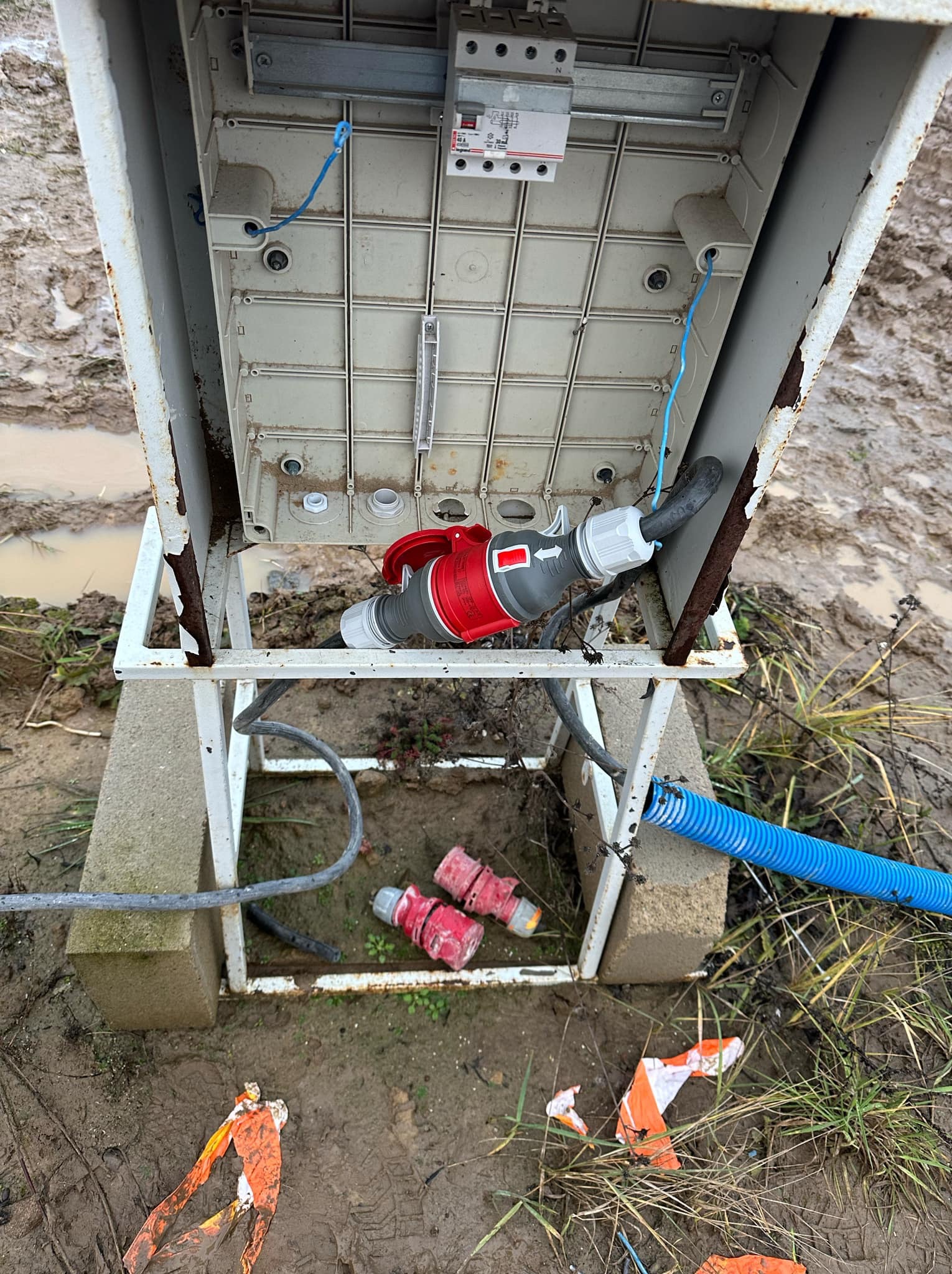 Skrzynka elektryczna z czerwonym gniazdem przemysłowym w błotnistym terenie, widoczne elementy instalacji i fragmenty taśmy ostrzegawczej.