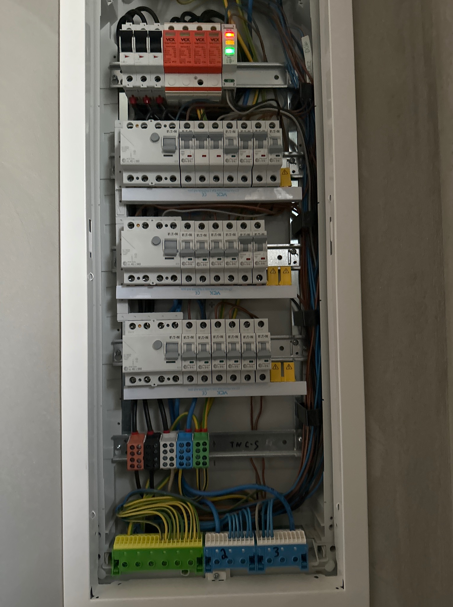 Otwarta skrzynka bezpiecznikowa z widocznymi bezpiecznikami, przewodami i listwami zaciskowymi, zamontowana na szarej ścianie.