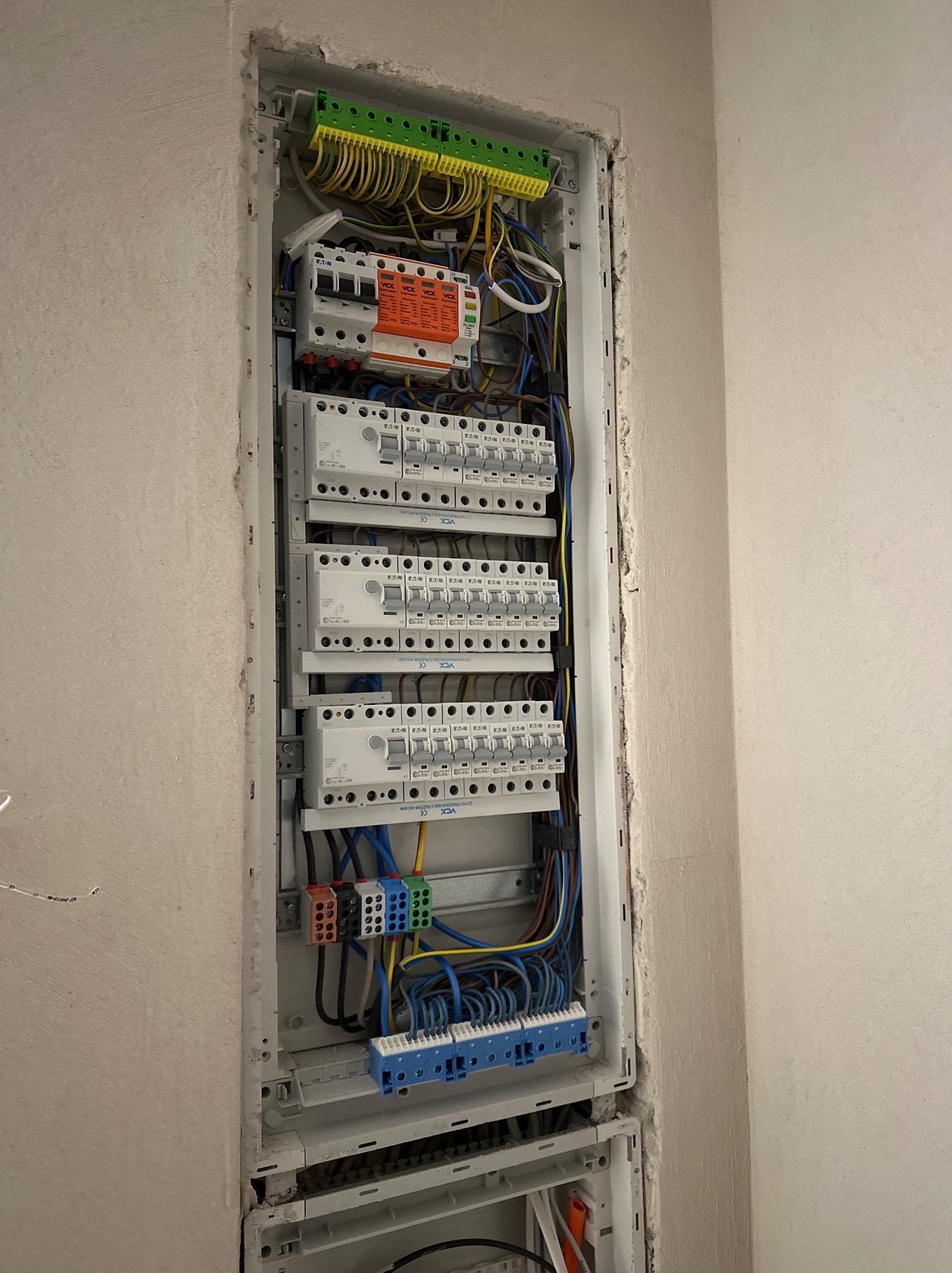 Rozdzielnica elektryczna z bezpiecznikami, zamontowana w ścianie, z widocznymi przewodami o różnych kolorach i złączkami, w surowym otoczeniu ściany przed wykończeniem.