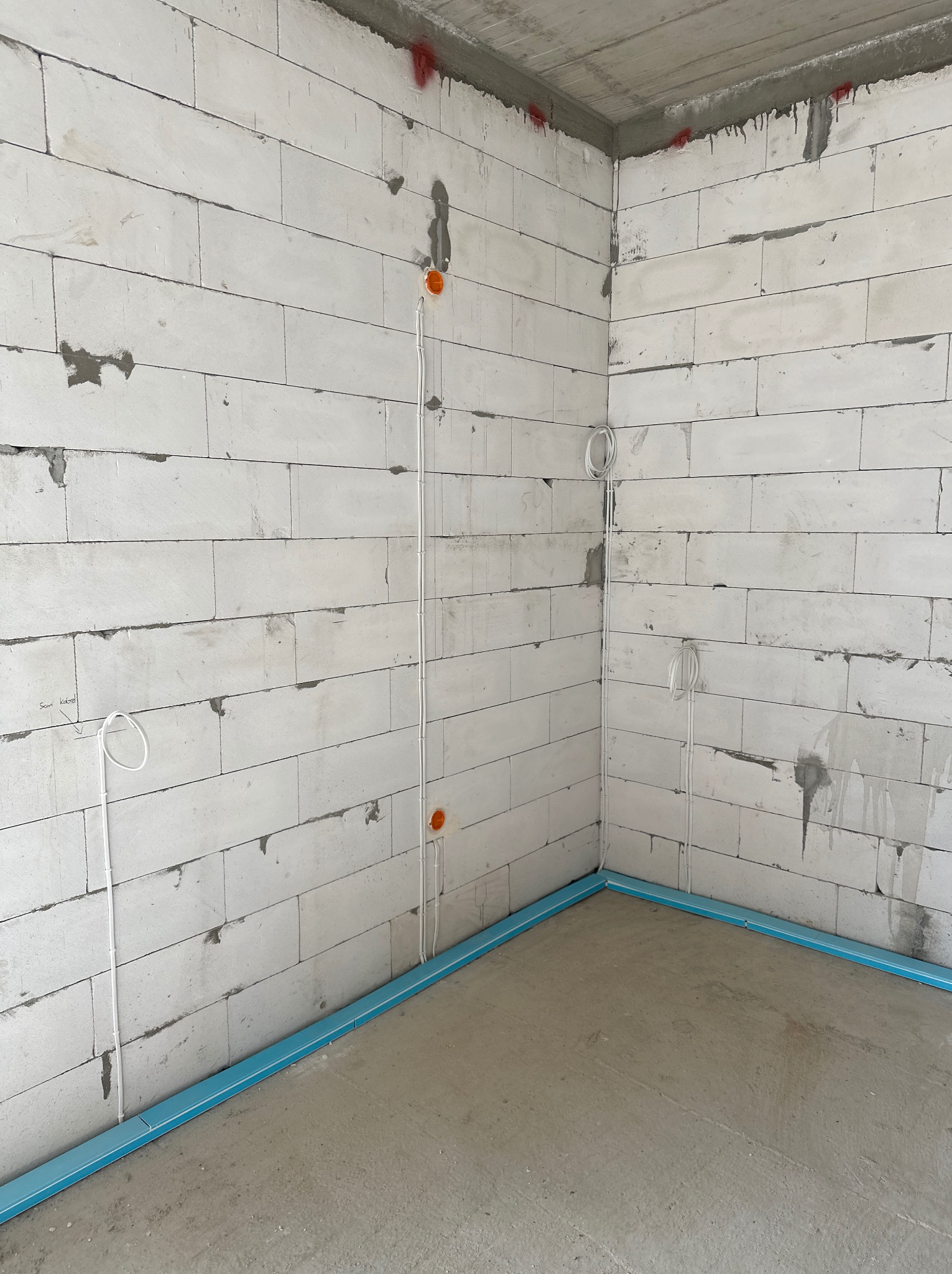 Surowy stan: Wnętrze w trakcie prac wykończeniowych, widoczne białe ściany z betonu komórkowego z poprowadzonymi przewodami elektrycznymi w peszlach, wystające pętle kabli, pomarańczowe puszki...