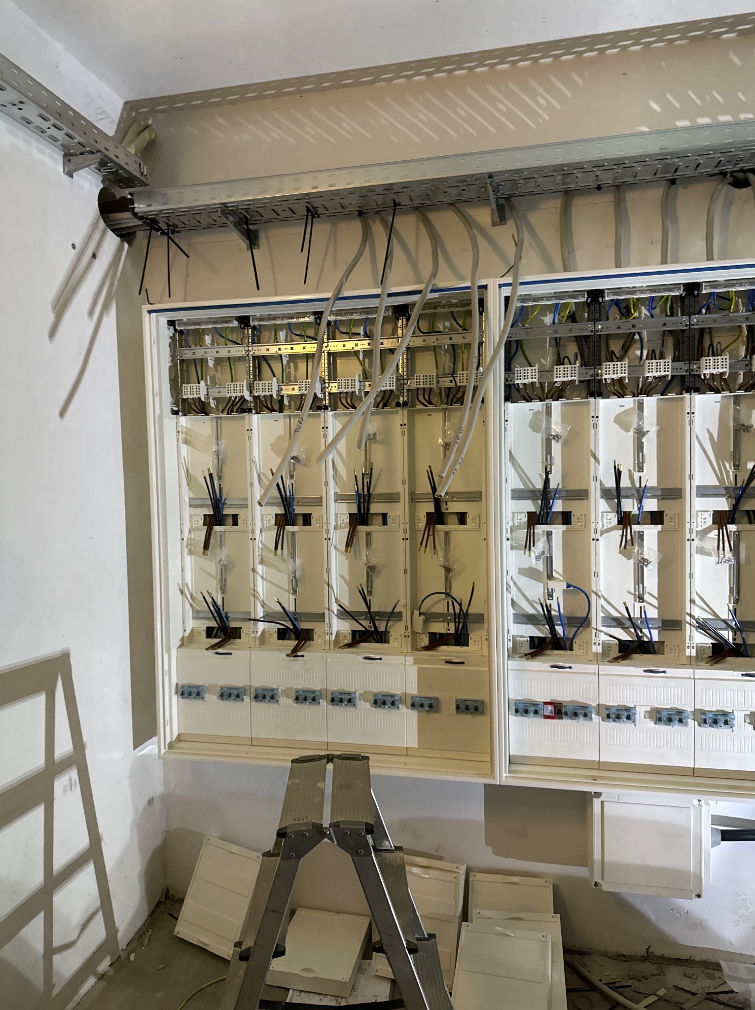 Rozdzielnica elektryczna w trakcie montażu, widoczne kable w peszlach, drabinki kablowe pod sufitem, drabina rozstawiona przed rozdzielnicą i pokrywy obudowy na podłodze.