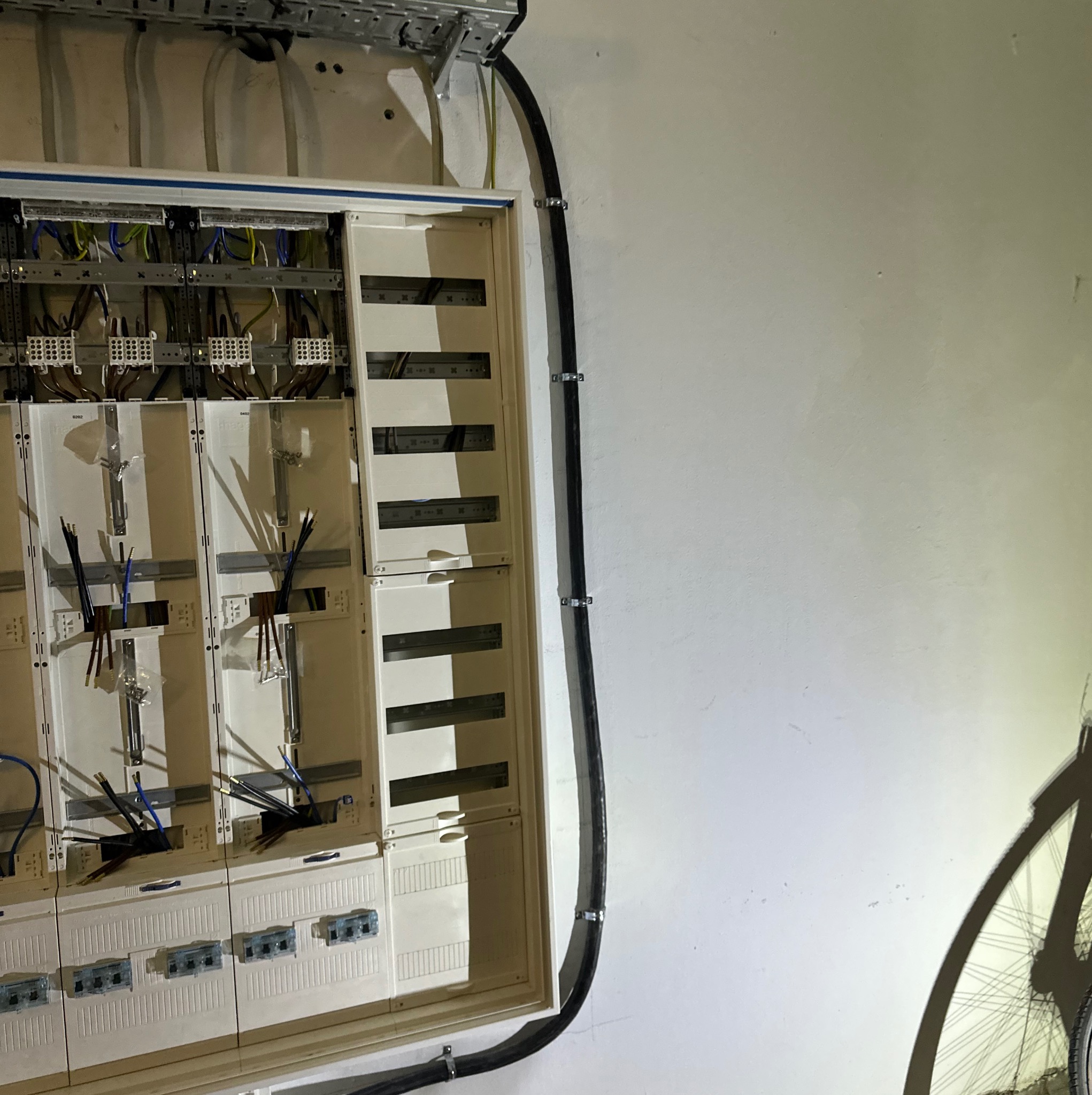 Rozdzielnica elektryczna w trakcie montażu, widoczne przewody, listwy zaciskowe i elementy mocujące, kabel elektryczny przytwierdzony do ściany, cień roweru.