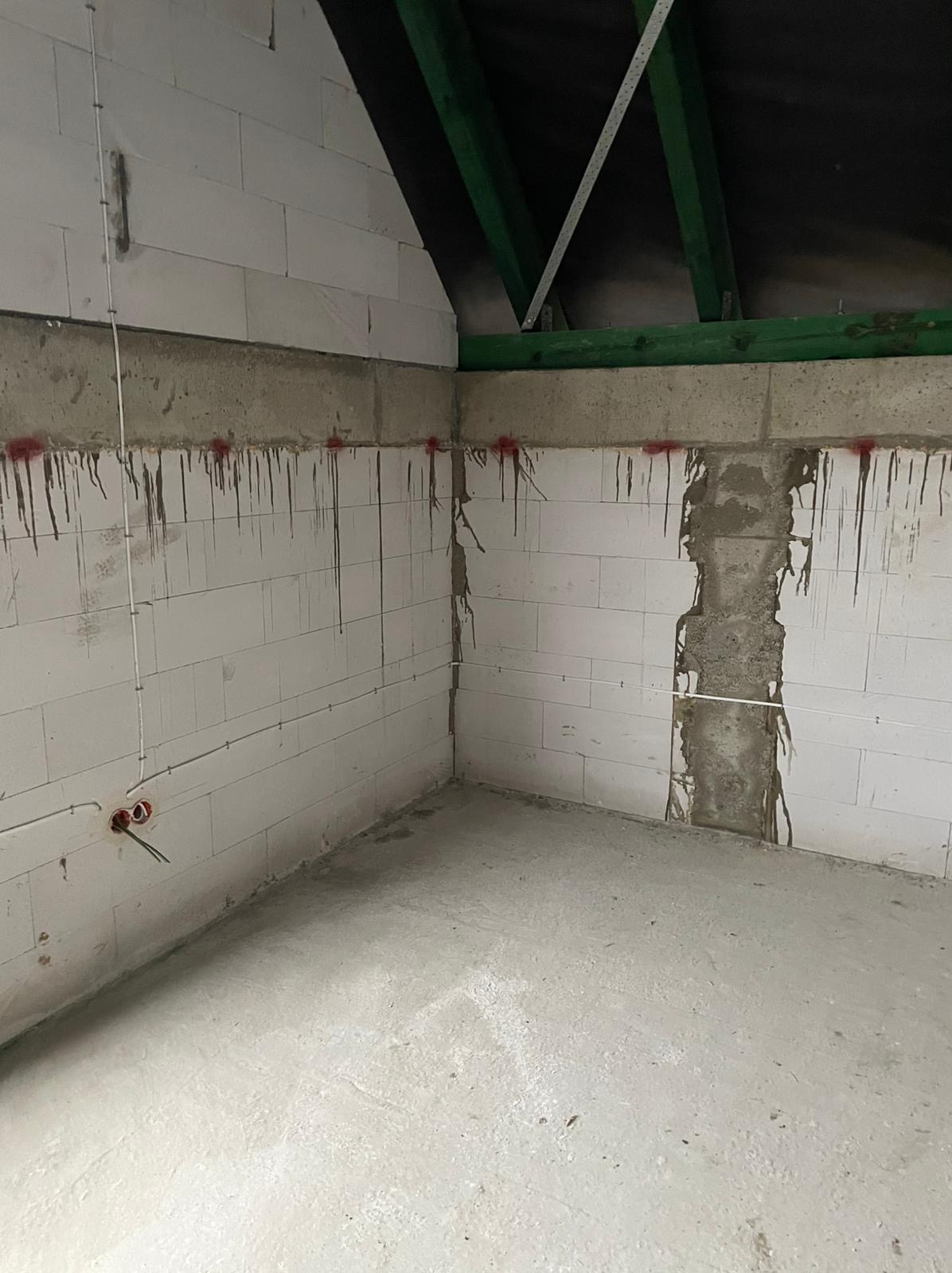 Surowe wnętrze poddasza w trakcie prac wykończeniowych: ściany z bloczków betonowych z widocznymi przewodami elektrycznymi i puszkami, ślady po wyznaczaniu punktów, surowy beton na podłodze...