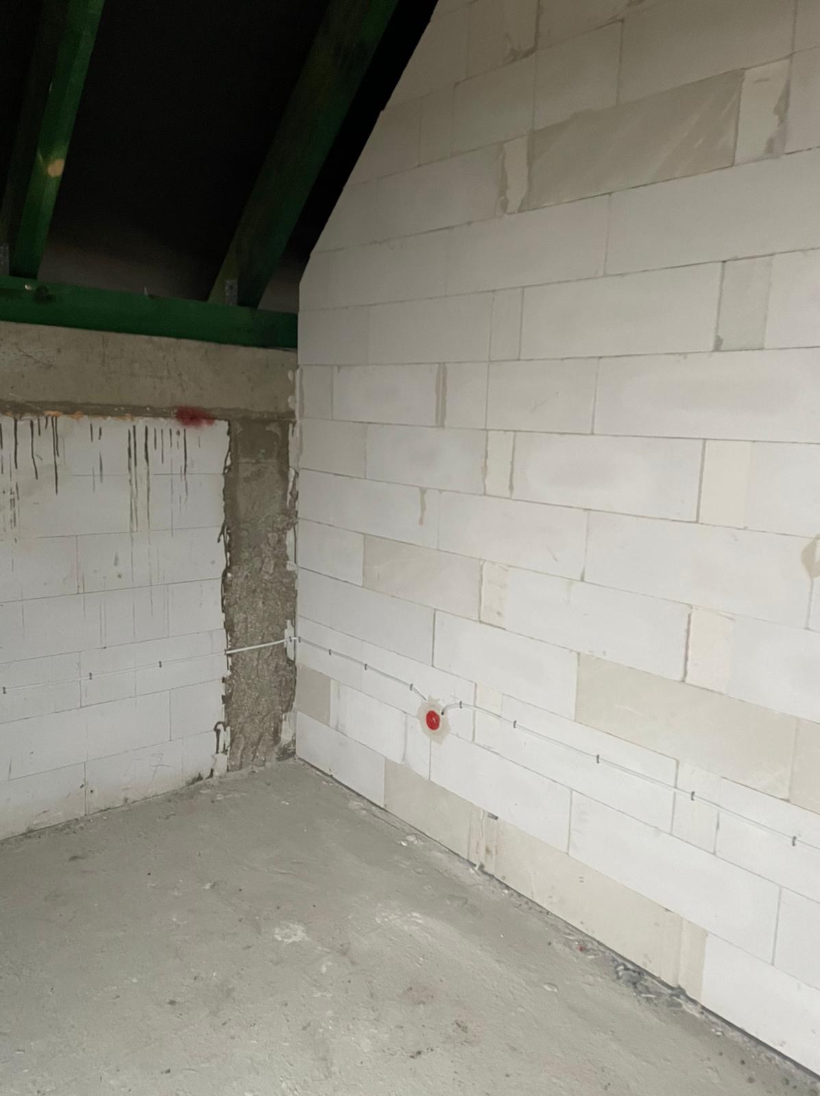 Surowy stan wnętrza z białymi bloczkami, widoczne przewody elektryczne prowadzone po ścianie z zamontowaną puszką instalacyjną z czerwoną osłoną, betonowa podłoga i fragment drewnianej konstrukcji...