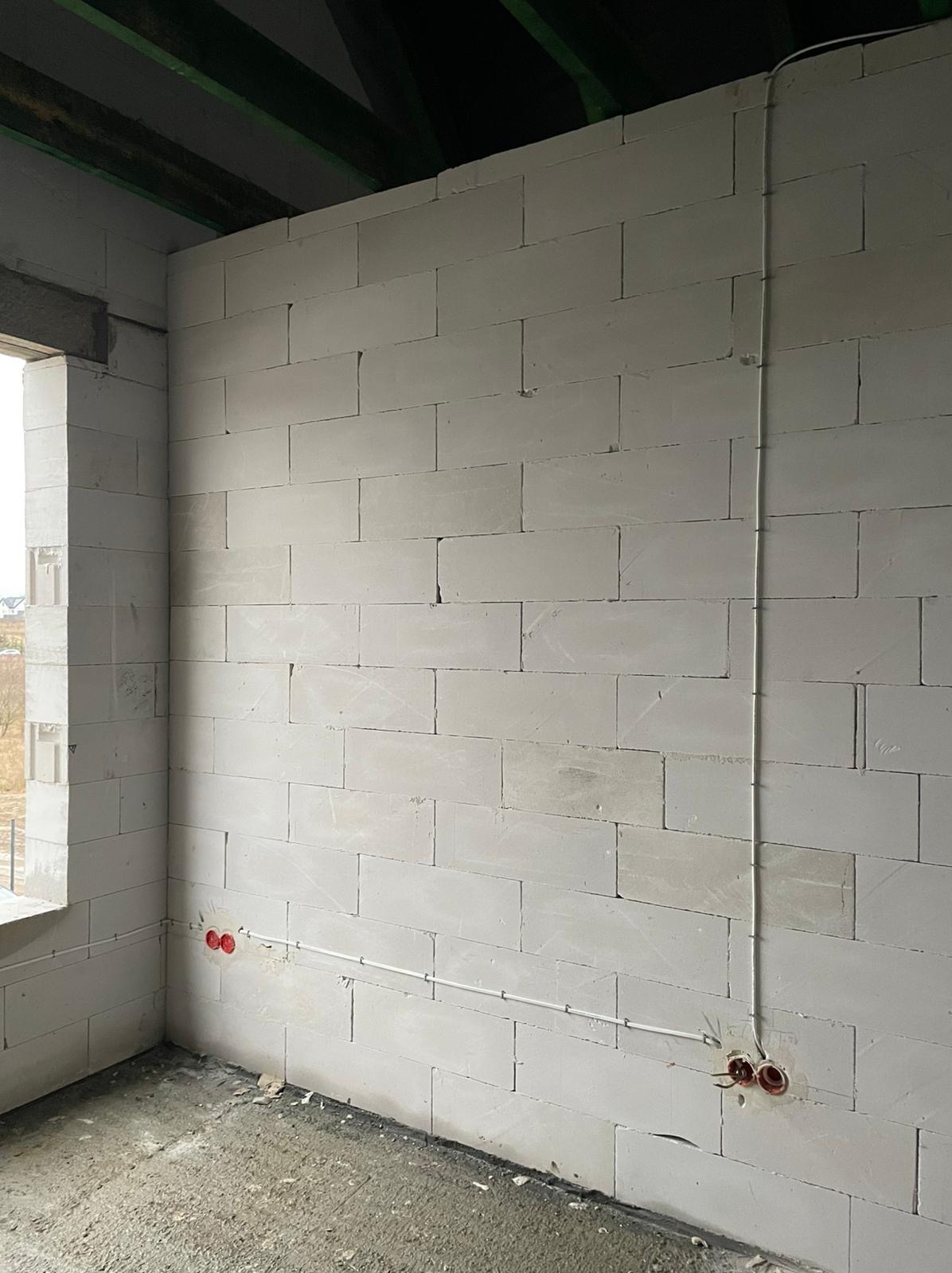 Surowa ściana z betonu komórkowego wewnątrz budynku w trakcie budowy, z widocznymi puszkami elektrycznymi i poprowadzonymi przewodami w białej rurce PCV.