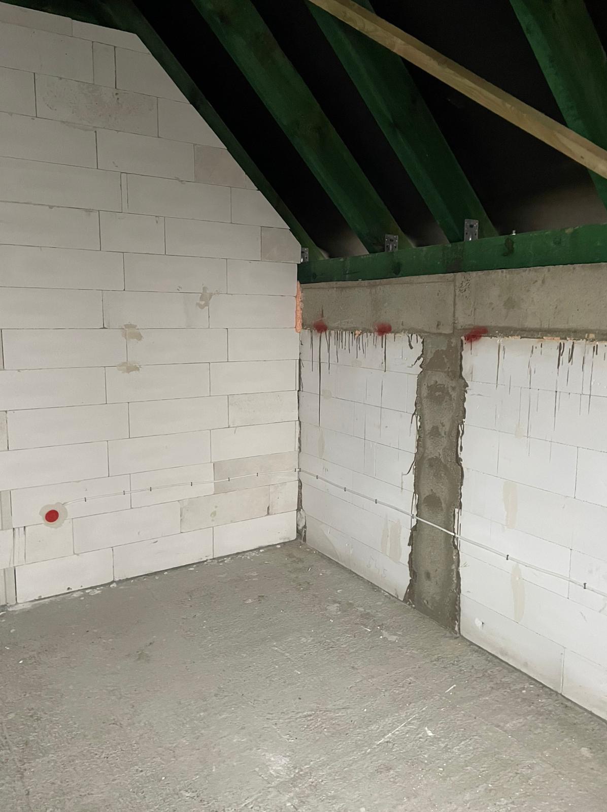 Surowe wnętrze poddasza z białymi ścianami z betonu komórkowego, zielonymi belkami stropowymi i betonową wylewką podłogową, widoczne przewody elektryczne w ścianach i czerwone oznaczenia na ścianach.