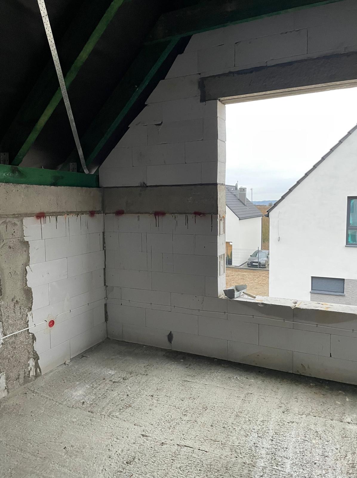 Surowy stan poddasza z widoczną instalacją elektryczną w puszce, ściany z białych bloczków, okno z widokiem na dom w tle.