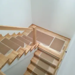 Jasne drewniane schody z balustradą, zabezpieczone kartonem, widok z góry. Minimalistyczny design, białe ściany. Detal wykończenia wnętrza.