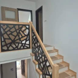 Nowoczesna balustrada drewniana z czarnym, geometrycznym wzorem, jasne drewno stopni schodów zabezpieczone kartonem, minimalistyczne wnętrze z drzwiami w tle.