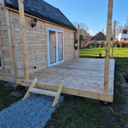 Usługi stolarskie Cezary - Drewniany taras z jasnego drewna przy domku w Oławie. Widoczne schody wejściowe i słupki podtrzymujące zadaszenie. Tło: trawnik i zabudowania.