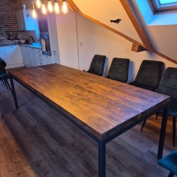 Usługi stolarskie Cezary - Stół z ciemnego drewna na metalowych nogach w nowoczesnym wnętrzu z kuchnią w tle. Widoczne krzesła i oświetlenie.