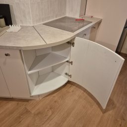Usługi stolarskie Cezary - Biała, narożna szafka kuchenna z otwartymi drzwiczkami, ukazująca półki wewnątrz. Blat w odcieniach szarości, płyta indukcyjna. Podłoga z paneli imitujących drewno.