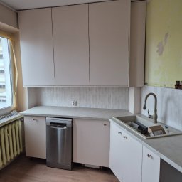 Usługi stolarskie Cezary - Jasna kuchnia z białymi szafkami, szarym blatem i srebrną zmywarką. Na ścianie białe, prostokątne płytki. Widoczne okno z grzejnikiem i fragment ściany w trakcie remontu.