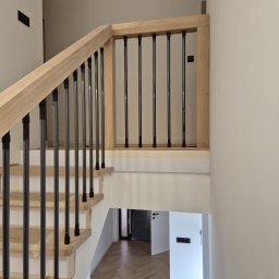 Usługi stolarskie Cezary - Jasne drewniane schody z czarnymi, metalowymi balustradami w nowoczesnym wnętrzu. Widoczny parkiet w jodełkę i białe ściany. Minimalistyczny design.