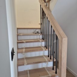 Usługi stolarskie Cezary - Jasne drewniane schody z czarnymi metalowymi balustradami w trakcie montażu, zabezpieczone kartonem i taśmą malarską. Widok z dołu na górę.