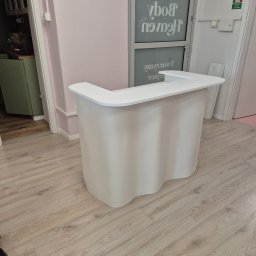 Usługi stolarskie Cezary - Biała lada recepcyjna o falistym kształcie na drewnianej podłodze, w tle ściana w odcieniach różu i drzwi z napisem. Minimalistyczny design.
