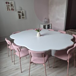 Usługi stolarskie Cezary - Owalny, biały stół z ośmioma różowymi krzesłami w pokoju o pastelowych ścianach. Na stole wazon z tulipanami i kartka. W tle półki z dekoracjami.