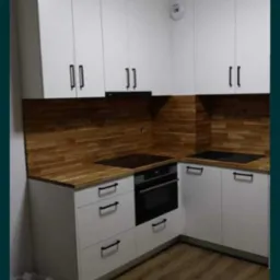 Kompaktowa kuchnia z białymi szafkami górnymi i dolnymi, czarnymi uchwytami, drewnianym blatem i panelem nad blatem w ciepłym odcieniu drewna, wyposażona w płytę indukcyjną i piekarnik.