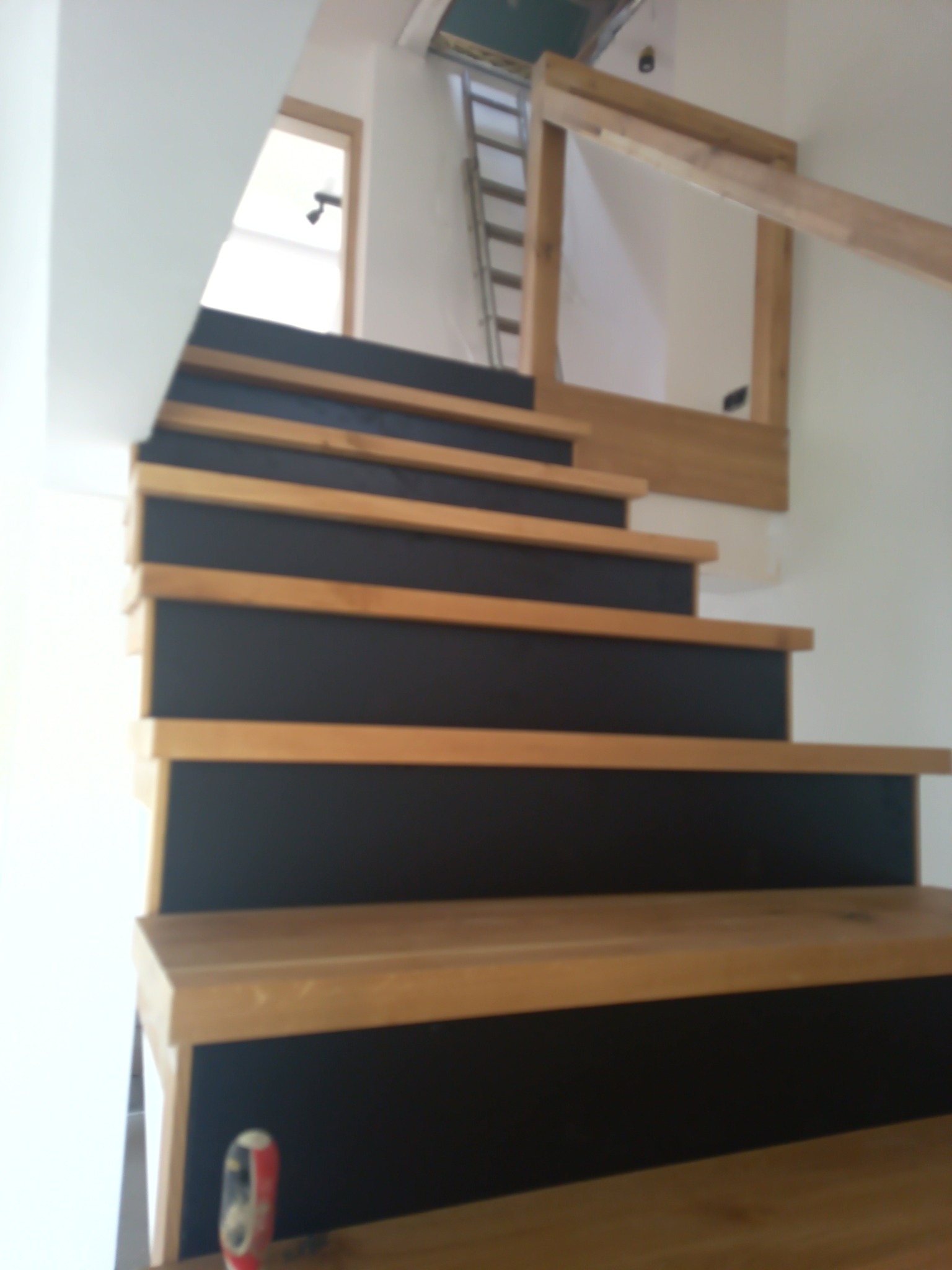 Schody drewniane z czarnymi podstopnicami, widoczne elementy konstrukcyjne i drabina w tle, perspektywa z dołu.