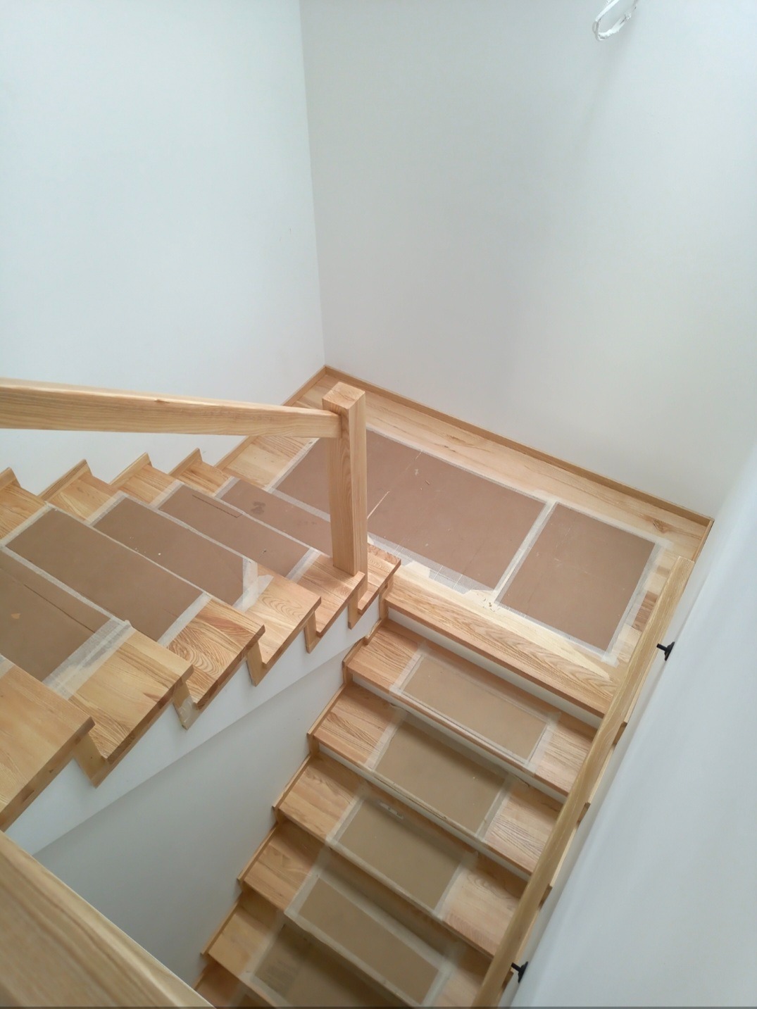 Jasne drewniane schody z balustradą, zabezpieczone kartonem, widok z góry. Minimalistyczny design, białe ściany. Detal wykończenia wnętrza.