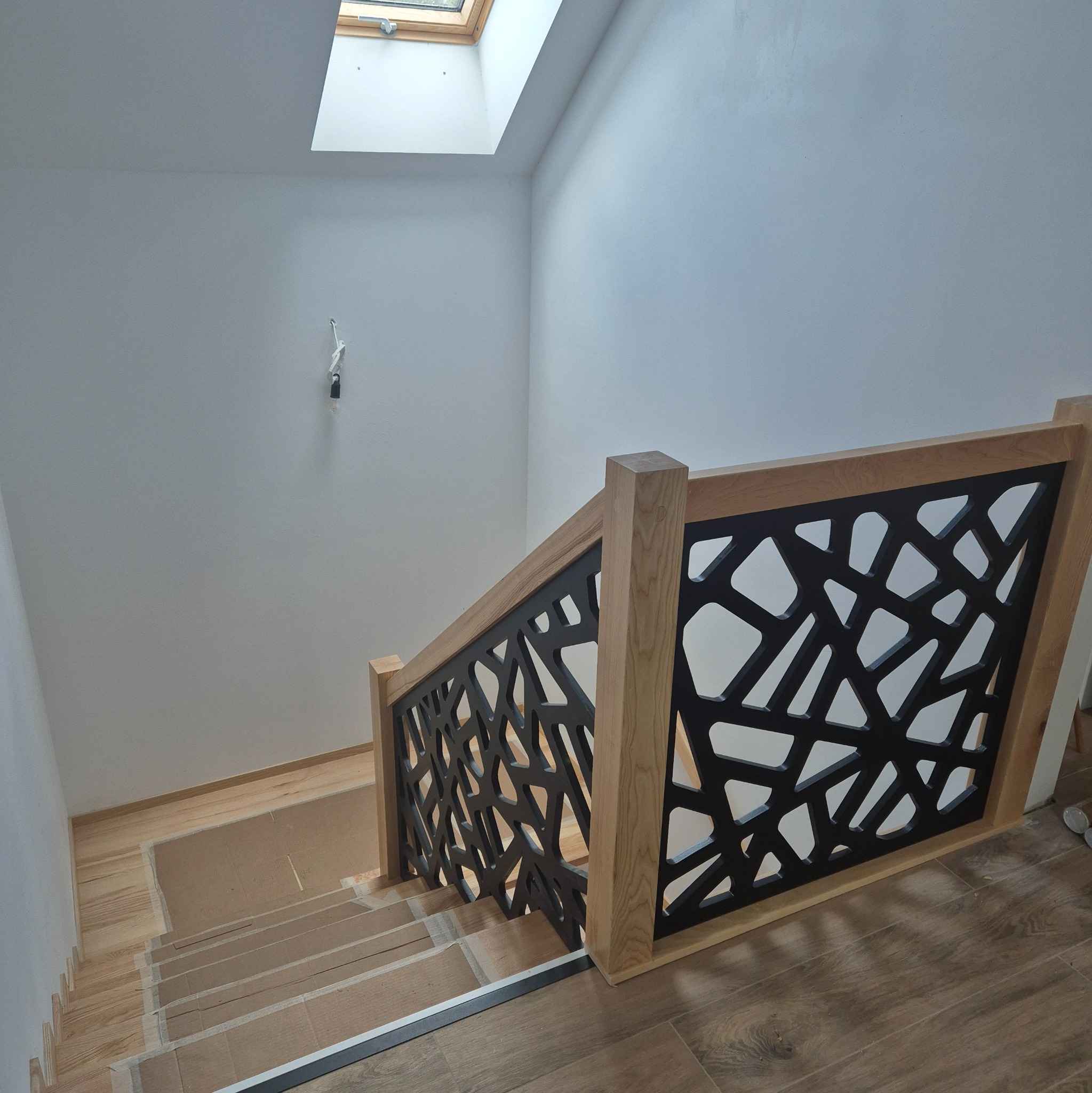 Nowoczesna balustrada schodowa z drewna i czarnego metalu o abstrakcyjnym wzorze, widok z góry na schody z drewnianymi stopniami i jasne ściany.