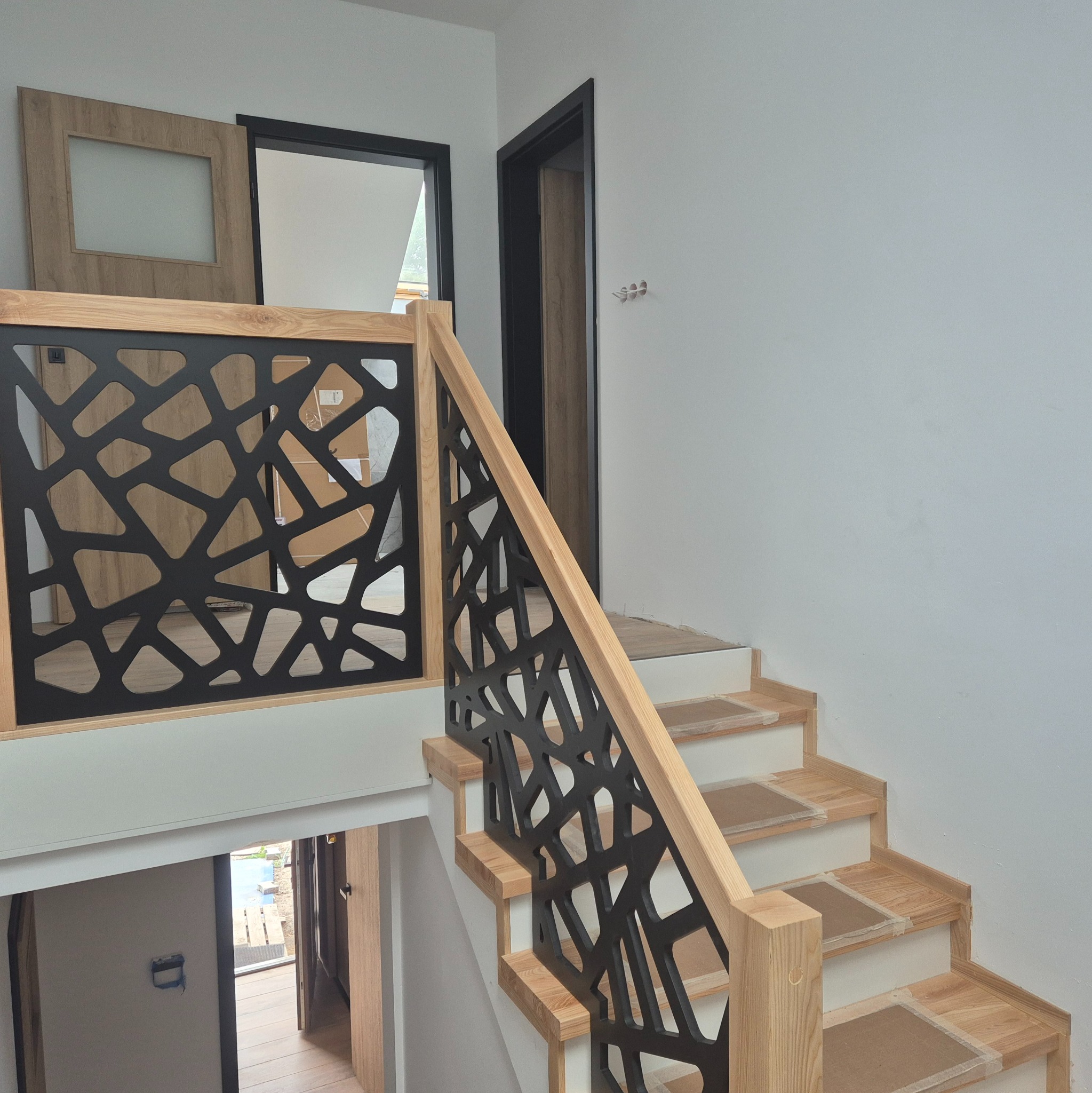 Nowoczesna balustrada drewniana z czarnym, geometrycznym wzorem, jasne drewno stopni schodów zabezpieczone kartonem, minimalistyczne wnętrze z drzwiami w tle.