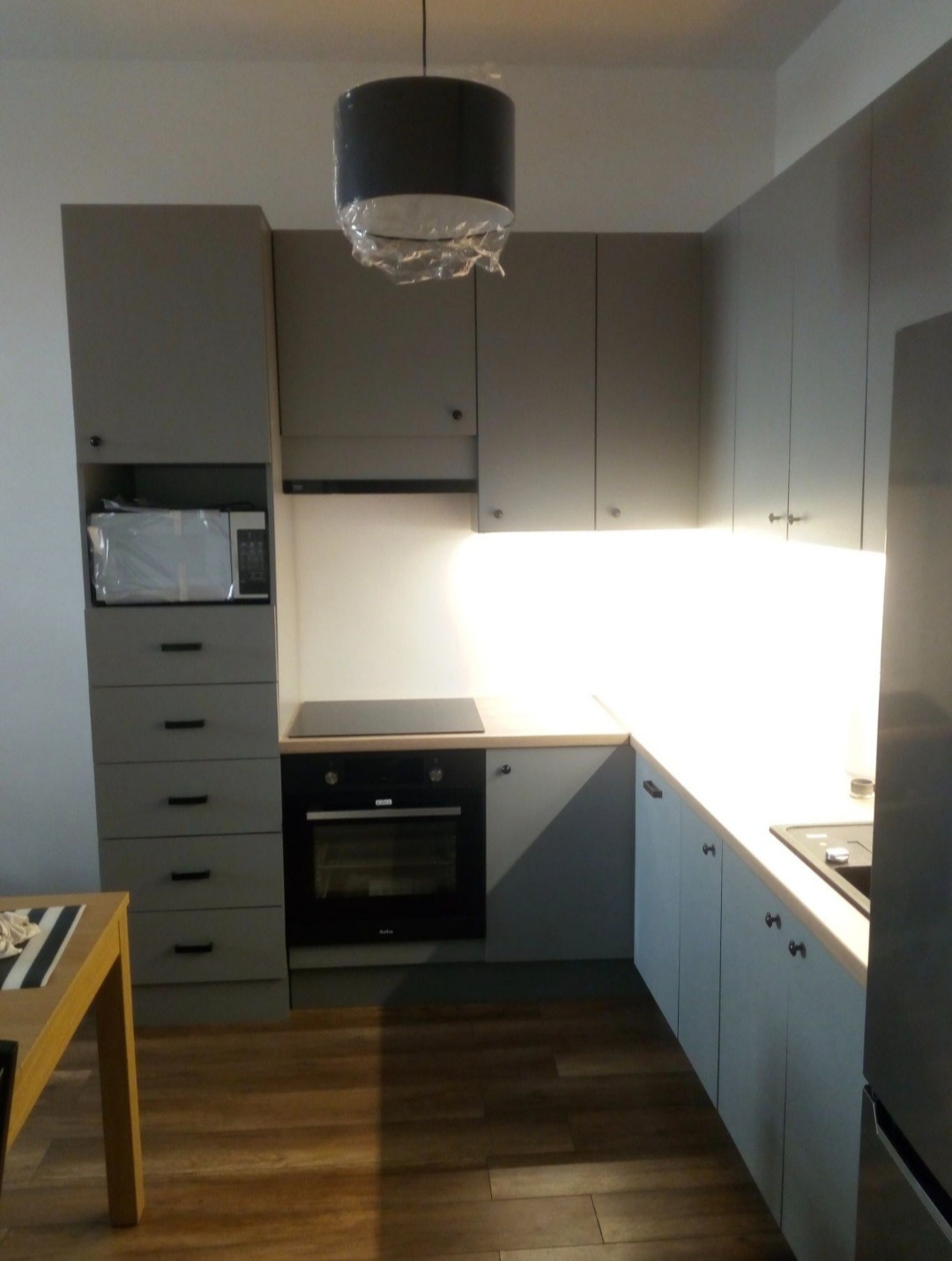 Szara zabudowa kuchenna z piekarnikiem i mikrofalówką. Nad blatem szafki wiszące z podświetleniem. Na suficie lampa z folią ochronną. Drewniana podłoga.