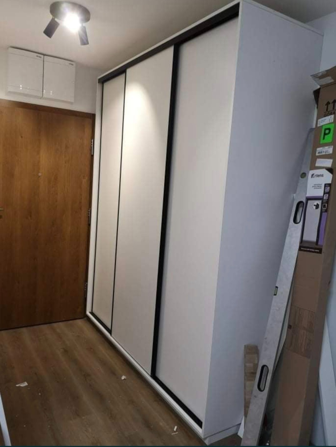 Nowoczesna szafa wnękowa z przesuwnymi drzwiami w kolorze białym i czarnymi obramowaniami, na tle drewnianej podłogi i drzwi.