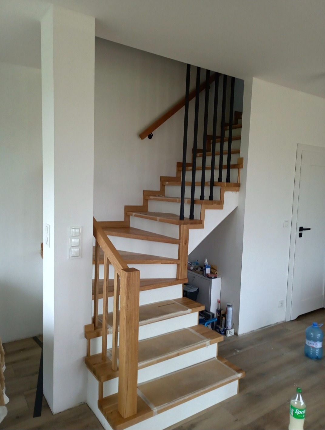Drewniane schody z czarnymi balustradami w jasnym wnętrzu, widok z dołu. Realizacja z Oławy. Stopnie zabezpieczone folią.
