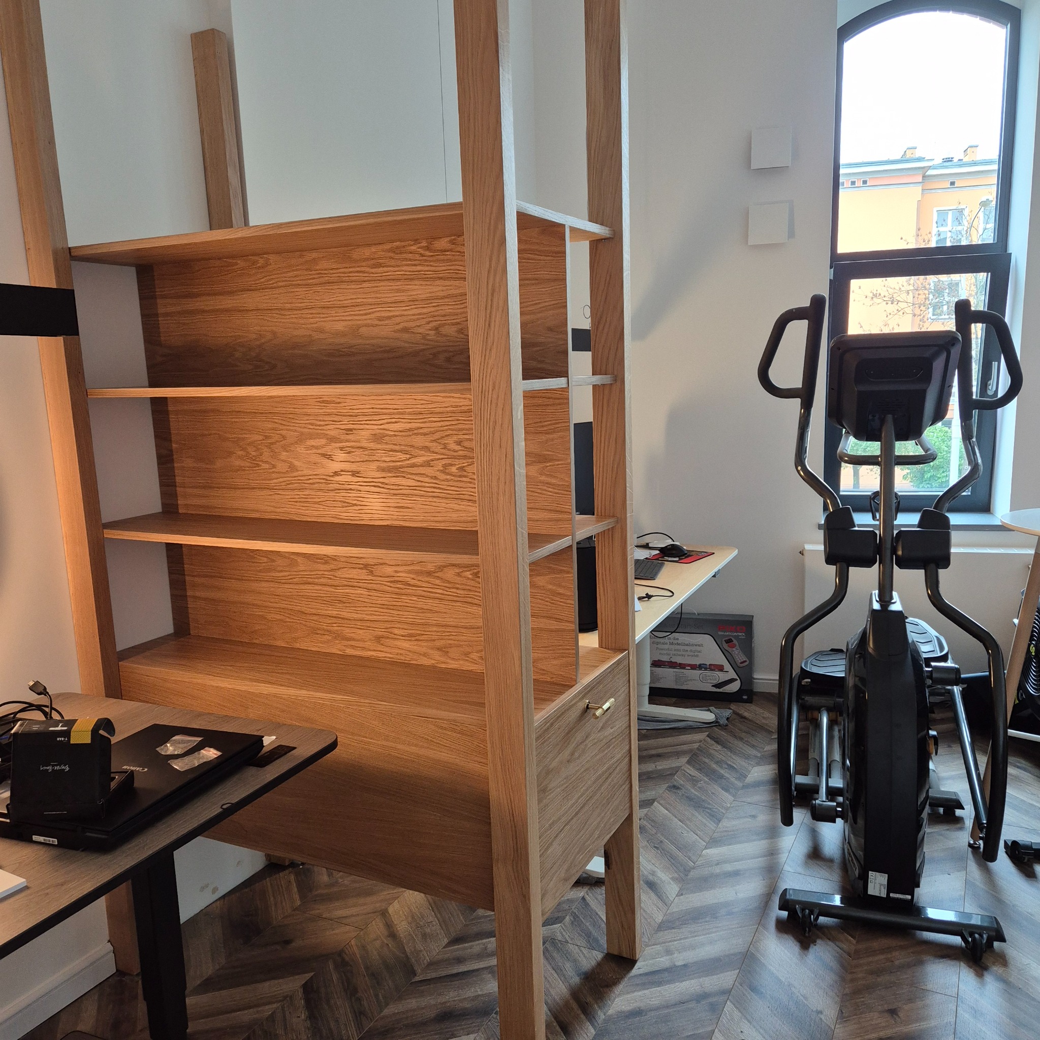 Drewniany regał z półkami i szufladą, wykonany na wymiar, z widocznym usłojeniem drewna, ustawiony w jasnym wnętrzu obok biurka i sprzętu do ćwiczeń.