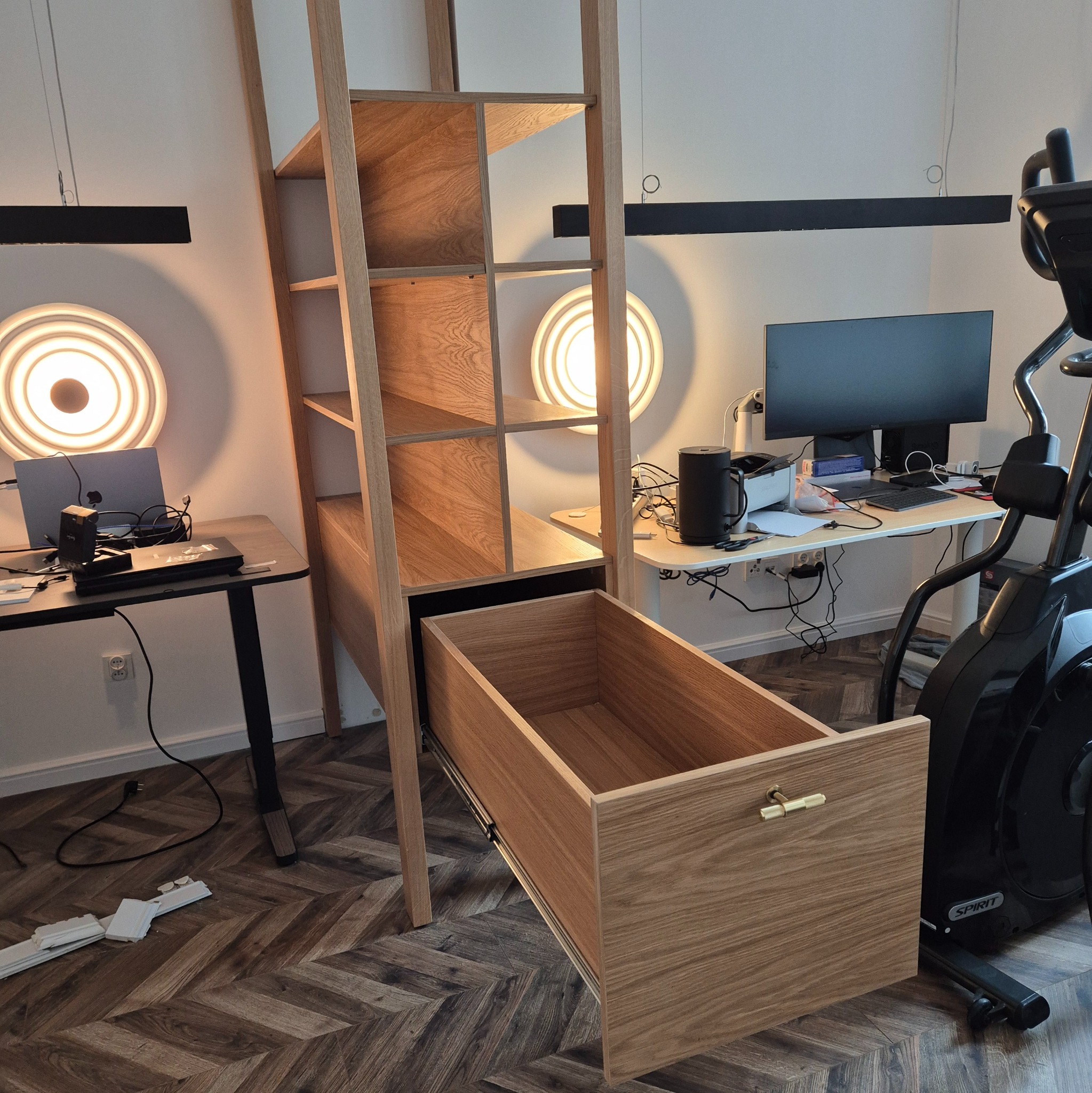 Drewniana szafa z otwartą szufladą na tle biurka z komputerem i lampami. Widoczny parkiet w jodełkę. Szafa z jasnego drewna, minimalistyczny design.