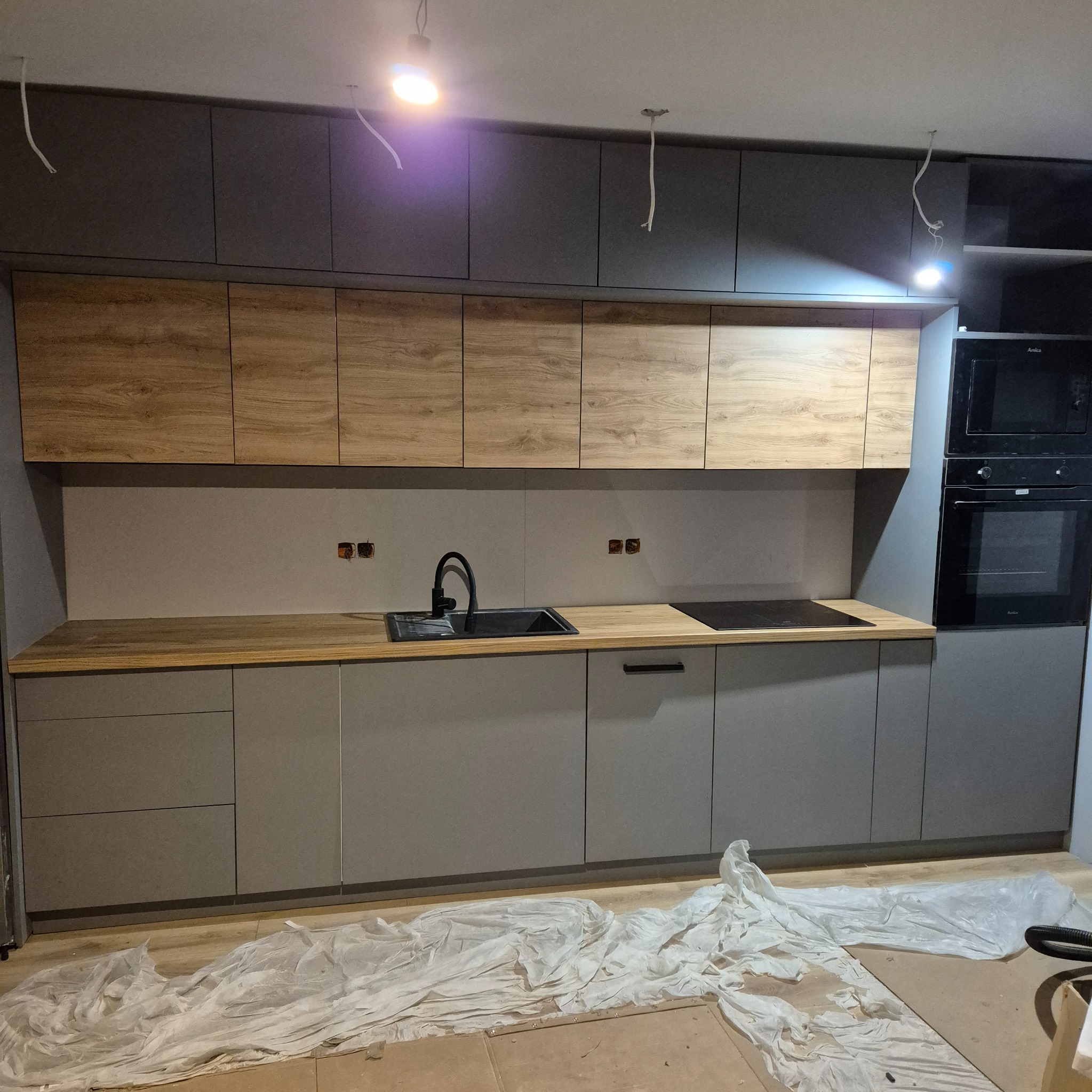 Szara kuchnia z drewnianymi szafkami górnymi, czarnym zlewem i płytą indukcyjną. Na podłodze folia ochronna. Widoczne niedokończone podłączenia elektryczne.