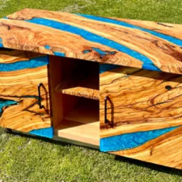 Niski regał z drewna o wyraźnym usłojeniu, wypełniony turkusową żywicą epoksydową, z otwartymi półkami i drzwiczkami z czarnymi uchwytami, na tle zielonej trawy.