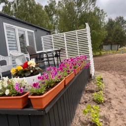 Grala Design Krystian Grala - Umeblowany taras z białym ażurowym ogrodzeniem i kwiatami w donicach, widok na ogród i drzewa w tle, szara elewacja domu.