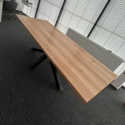 Rozkładany stół z jasnego drewna na czarnych, metalowych nogach w nowoczesnym wnętrzu z żaluzjami. Widoczny fragment blatu i nóg.