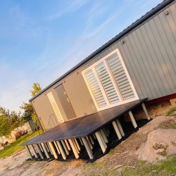 Grala Design Krystian Grala - Nowoczesny, szary domek z ciemnym tarasem na rurkowych podporach, widziany z boku pod kątem, na tle błękitnego nieba. Minimalistyczny design.