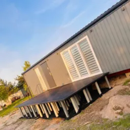 Nowoczesny, szary domek z ciemnym tarasem na rurkowych podporach, widziany z boku pod kątem, na tle błękitnego nieba. Minimalistyczny design.