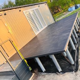 Grala Design Krystian Grala - Nowoczesny taras kompozytowy przy budynku z szarymi podporami, widok z boku, z zieloną miotłą opartą o schody wejściowe.