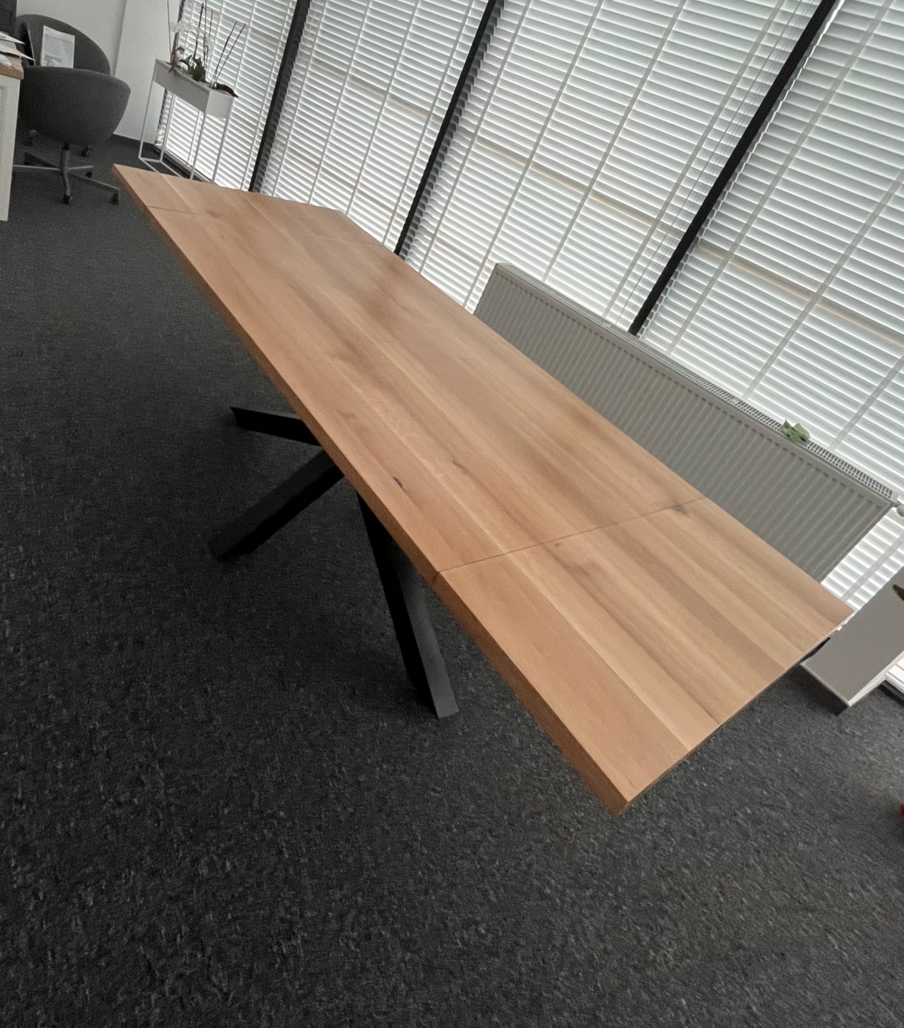 Rozkładany stół z jasnego drewna na czarnych, metalowych nogach w nowoczesnym wnętrzu z żaluzjami. Widoczny fragment blatu i nóg.