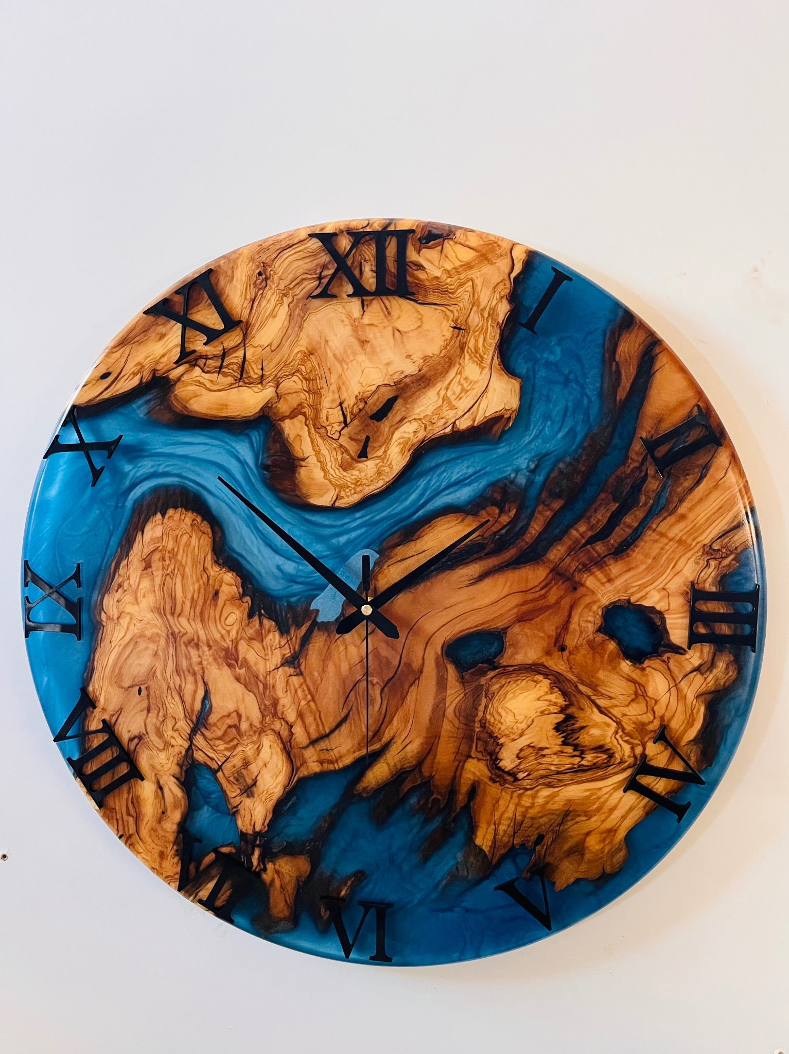 Okrągły zegar ścienny z drewna oliwkowego i żywicy epoksydowej w kolorze błękitnym, z czarnymi rzymskimi cyframi, wskazujący godzinę.