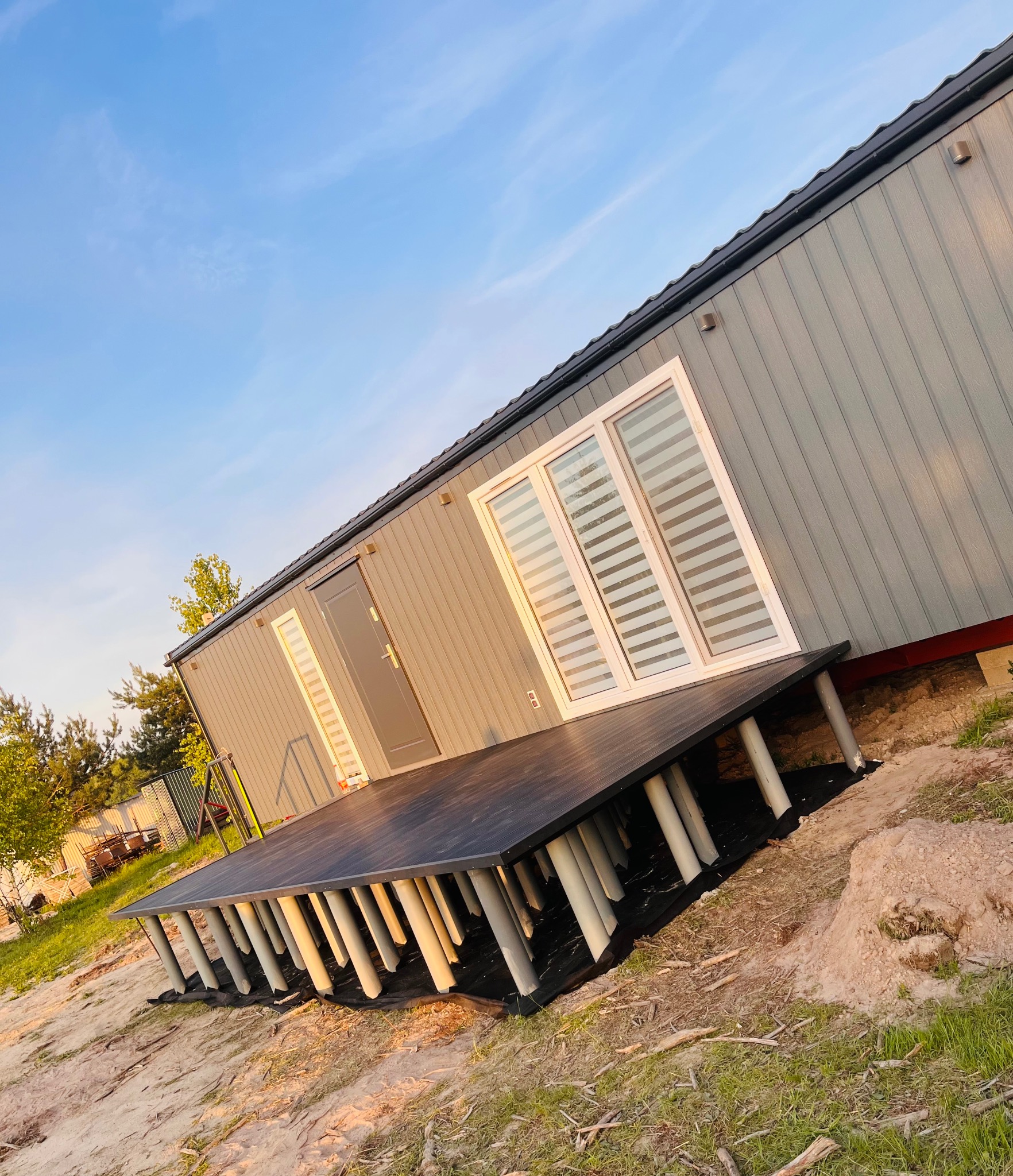 Nowoczesny, szary domek z ciemnym tarasem na rurkowych podporach, widziany z boku pod kątem, na tle błękitnego nieba. Minimalistyczny design.