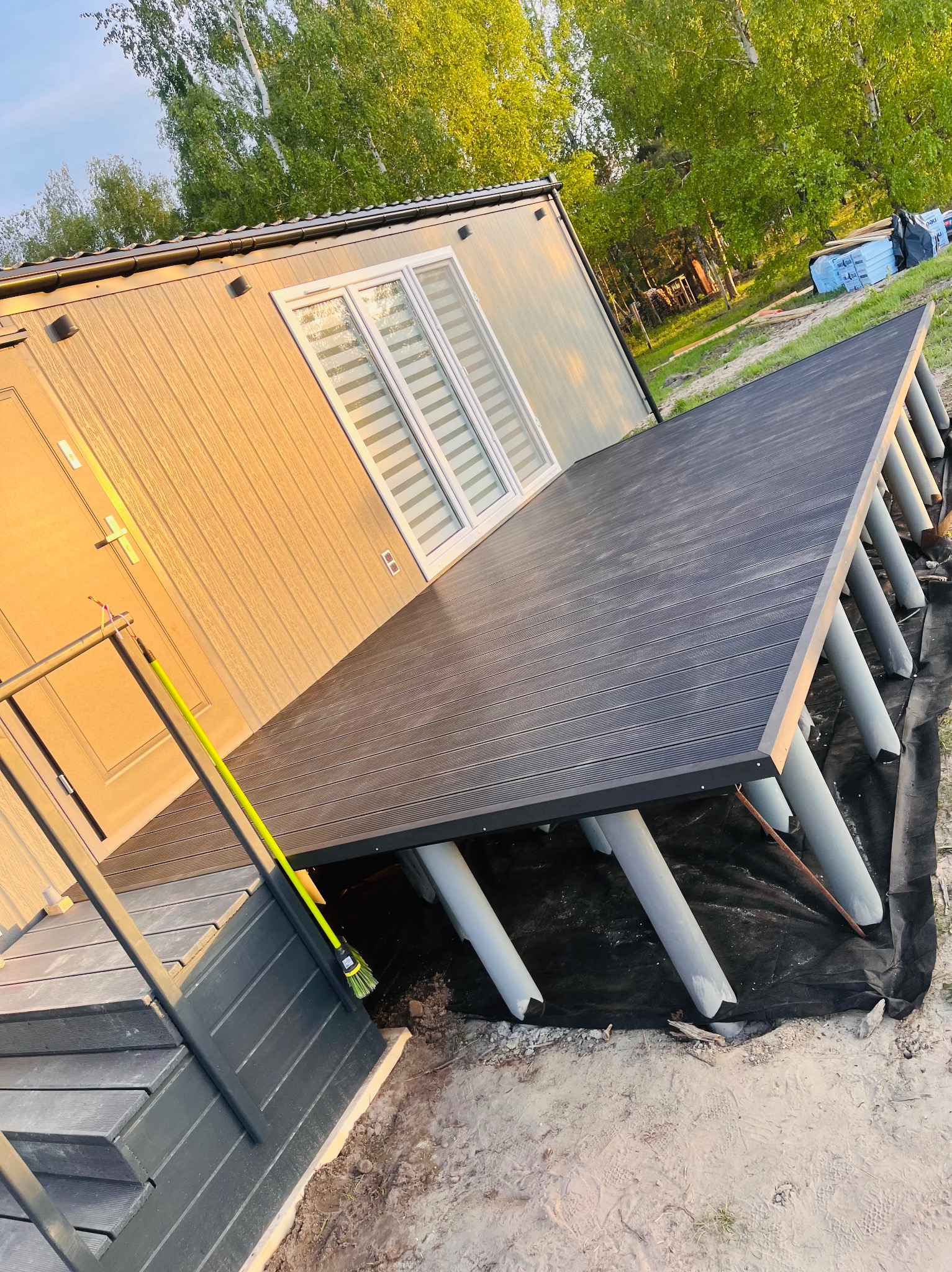 Nowoczesny taras kompozytowy przy budynku z szarymi podporami, widok z boku, z zieloną miotłą opartą o schody wejściowe.