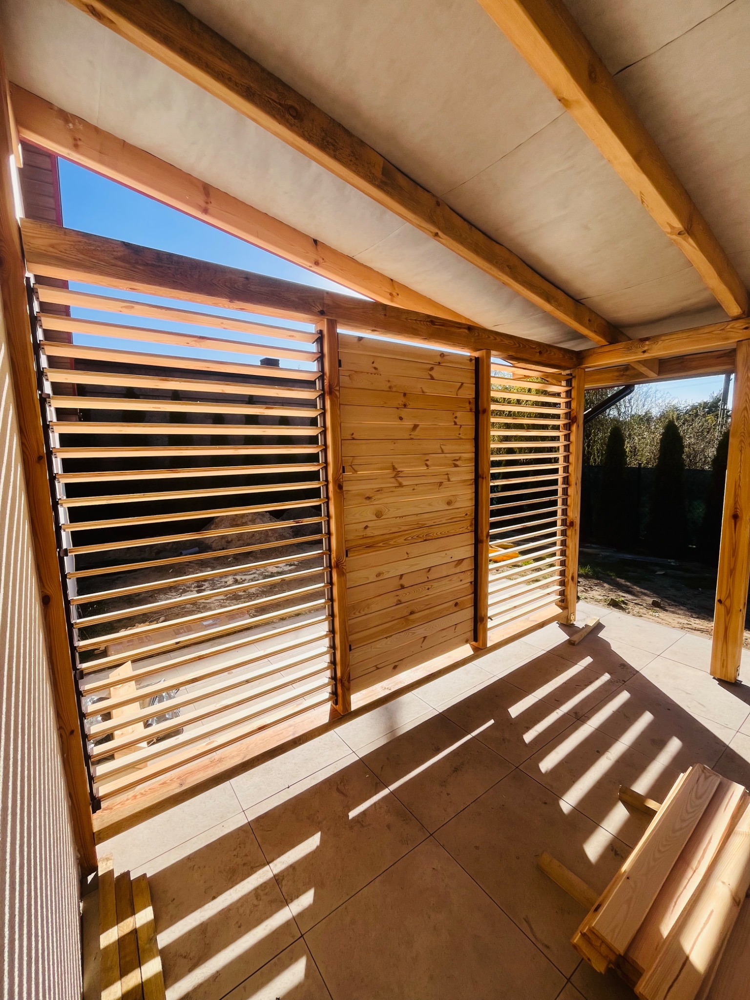 Drewniana altana z lamelami w słoneczny dzień. Widoczne elementy konstrukcyjne i cień padający na podłogę. Częściowo ukończona konstrukcja.