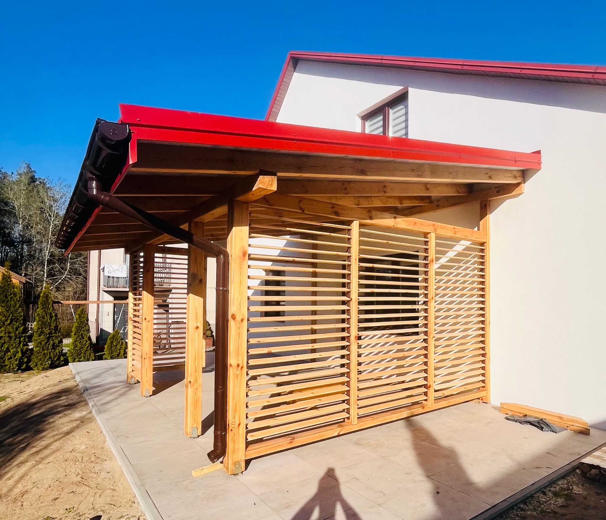 Drewniana altana z lamelami i czerwonym dachem, przylegająca do białego budynku. Widoczne detale konstrukcyjne i elementy wykończeniowe.