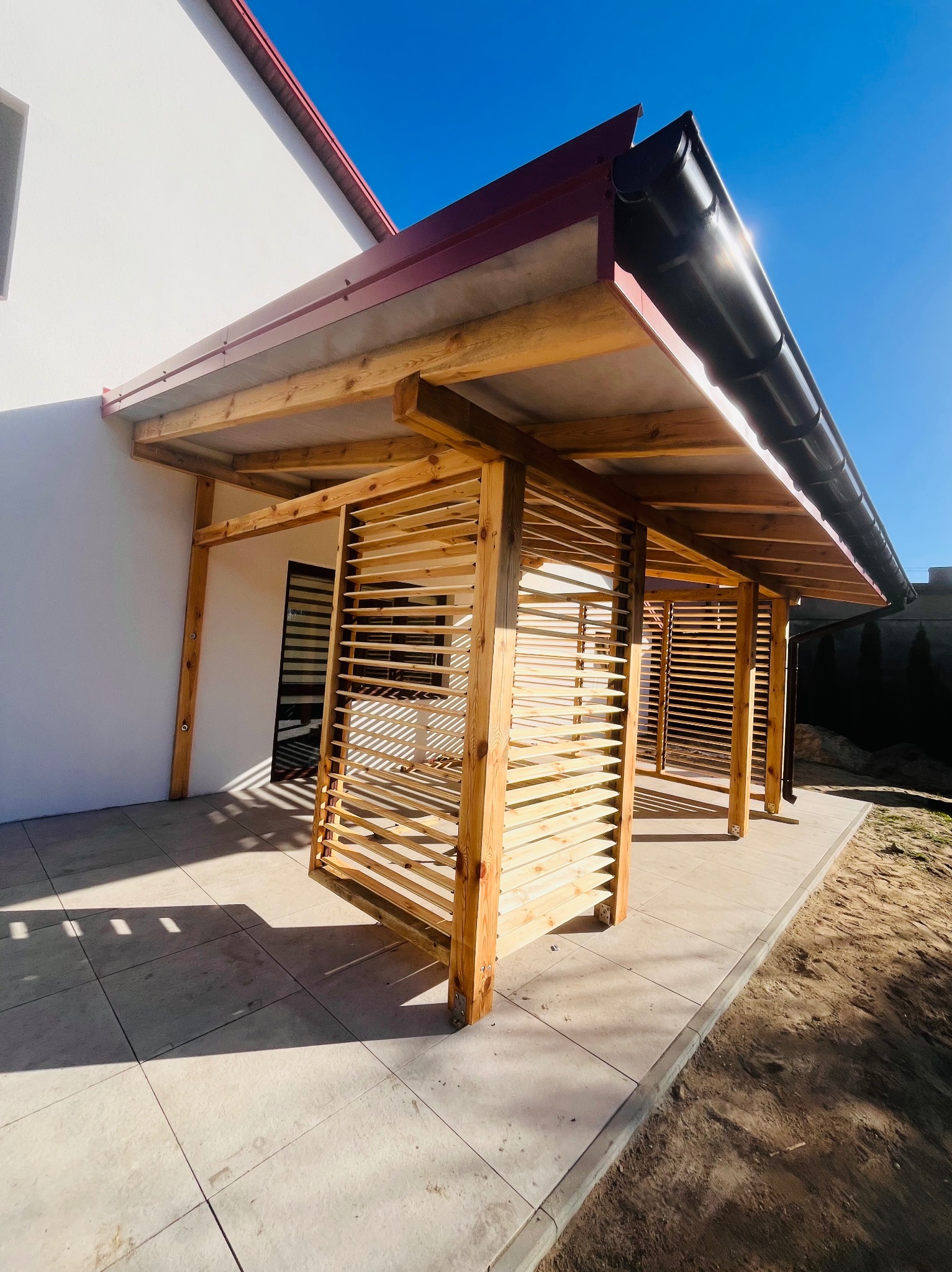 Drewniana konstrukcja z żaluzjami przy budynku z czerwoną rynną i jasną elewacją, jasne płytki na ziemi i błękitne niebo w tle. Kontrastowe oświetlenie.
